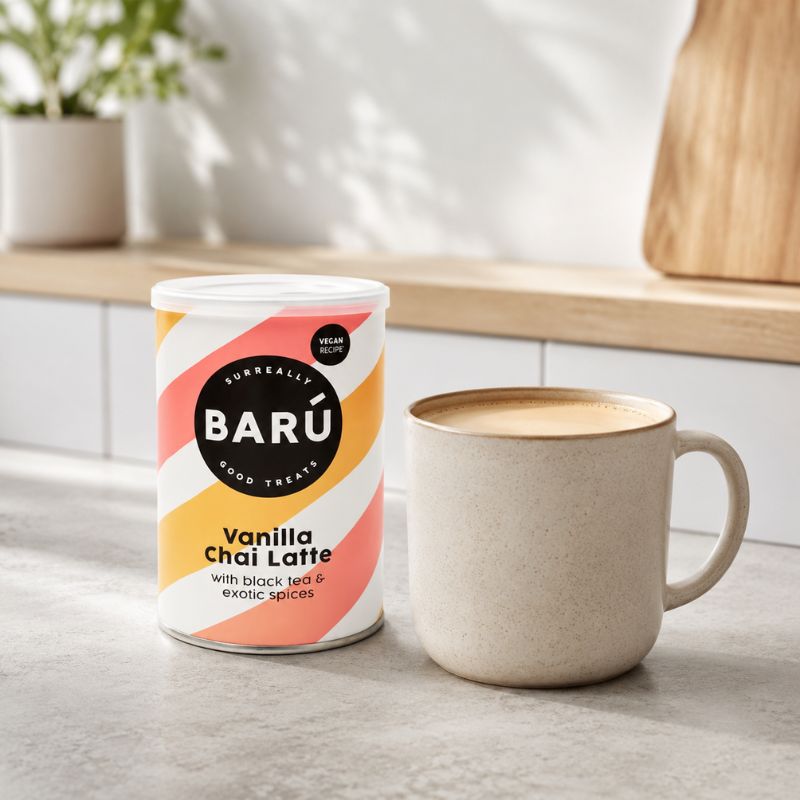 Barú Vanilla Chai Latte 250 g ved siden av et keramikkrus med chai latte på en lys kjøkkenbenk.