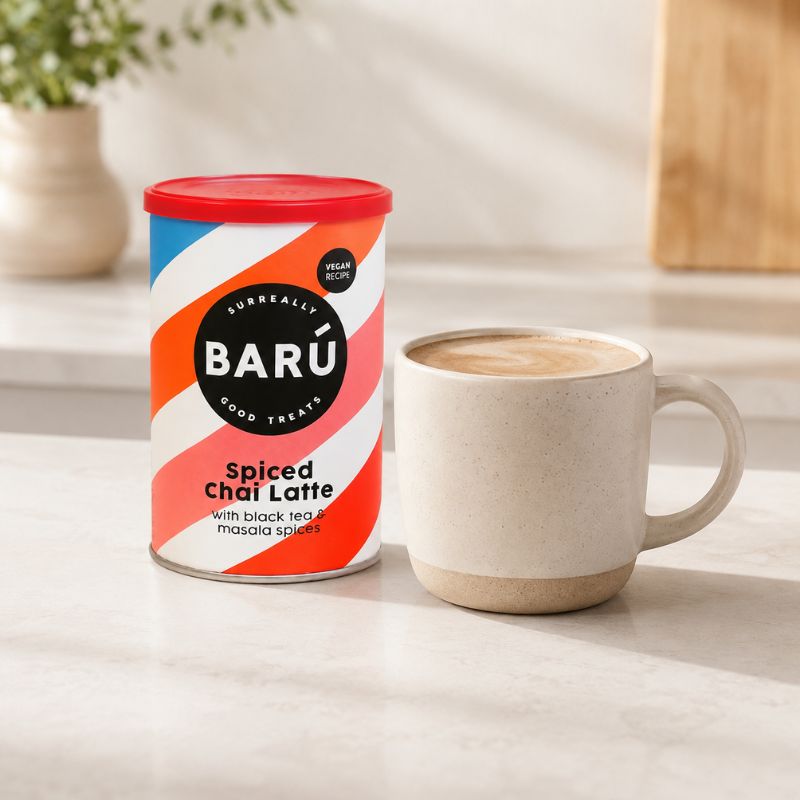 Barú Spiced Chai Latte 250 g ved siden av et keramikkrus med chai latte på en lys kjøkkenbenk.